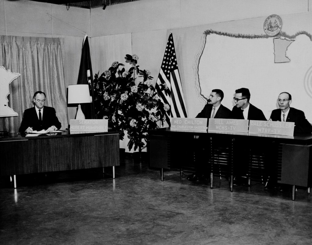 This photo shows Senator Hubert Humphrey seated at the left of the photo behind a desk as part of the Kennedy-Humphrey debate that took place in Charleston, 1960. Three news reporters sit behind name plaques at the righthand side. 