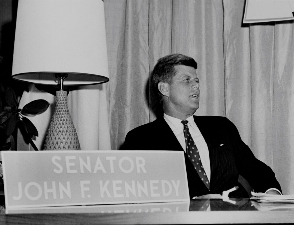 Senator John F. Kennedy sits behind a desk, with a lamp to his left and a name plaque in the foreground.