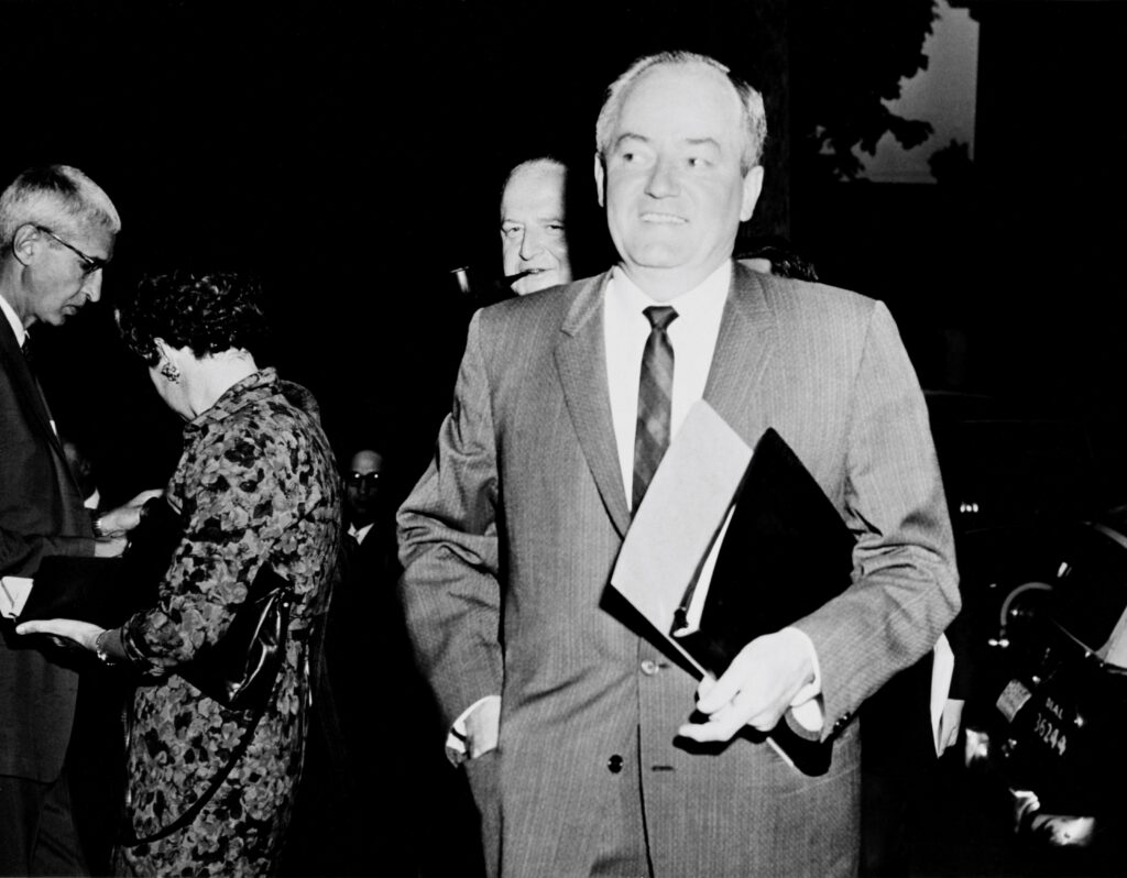 Senator Humphrey walks towards to camera. He is holding some files under his arm. a few people can be seen in the background. 