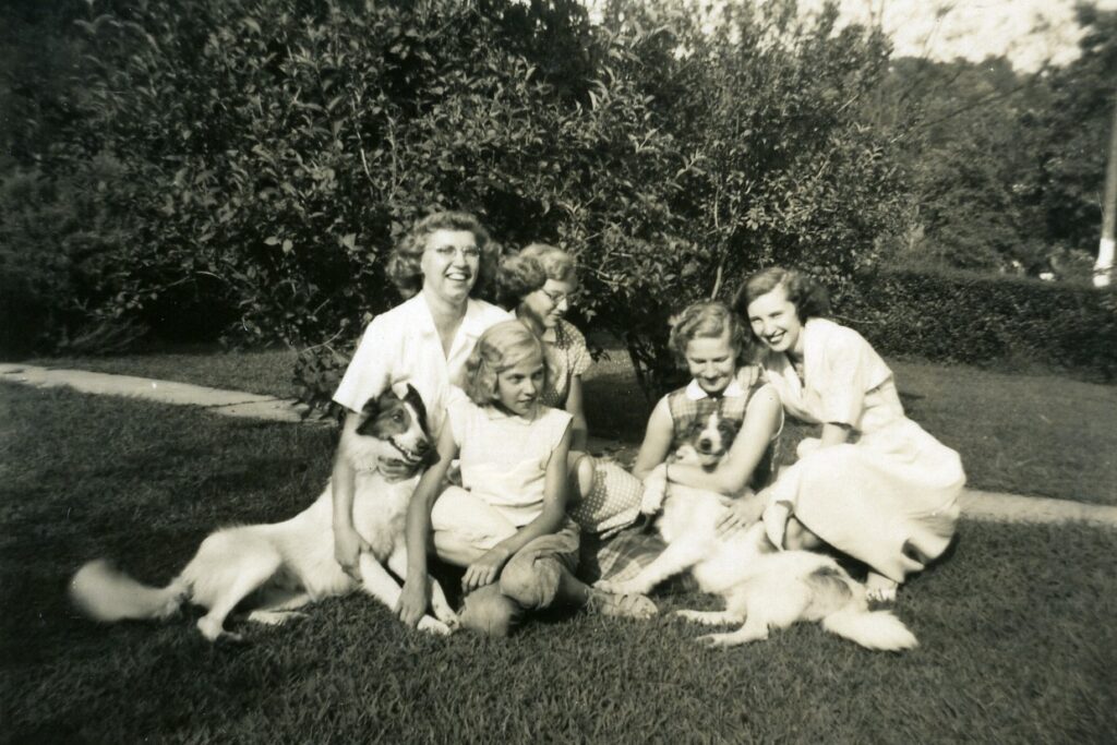 A black and white photo of five sisters seated on the lawn. They appear to be laughing, two dogs have joined them for the photo.