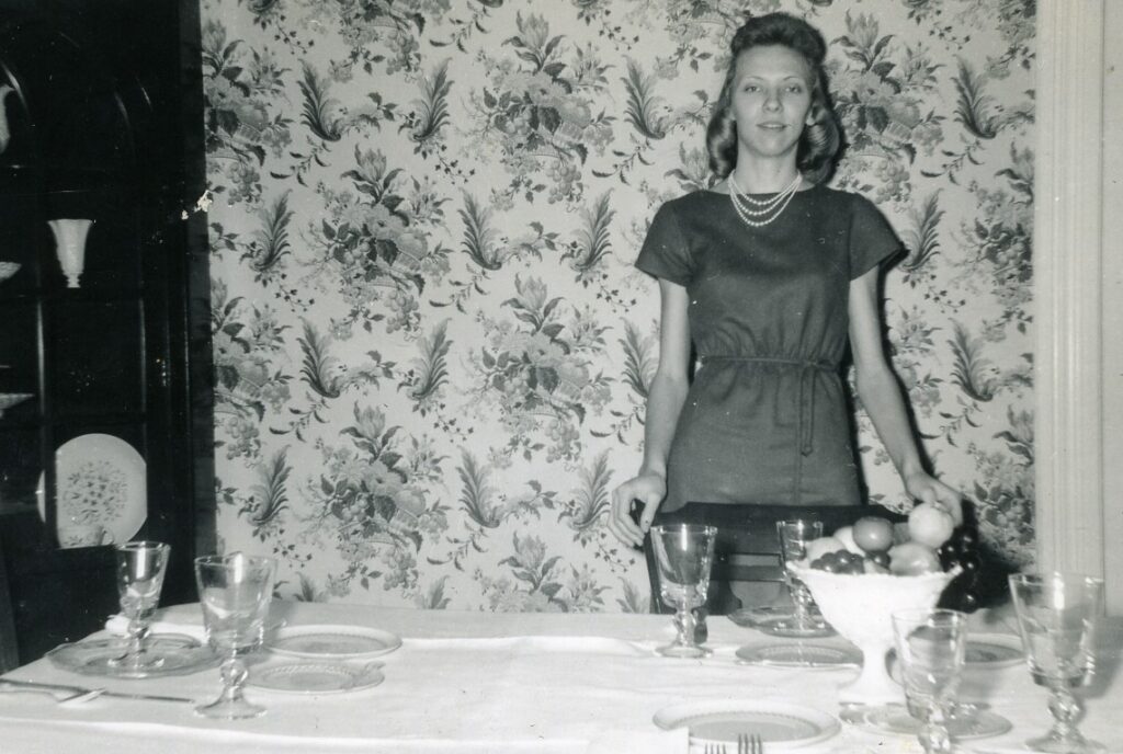 Frances Peters stands behind a table, an empty place setting and a bow of fruit sit on the table in the foreground.