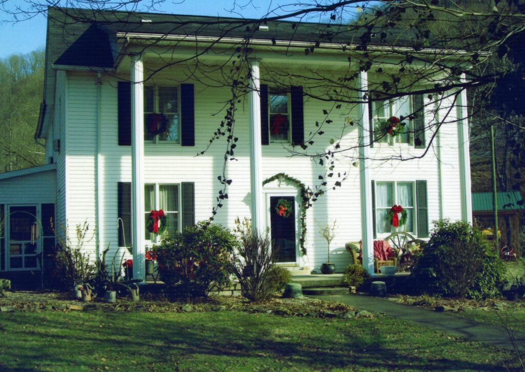 A photo of a large two-story historic home with four columns across the front. There are wreathes with red bows on each window, as if decorated for the Christmas season. 