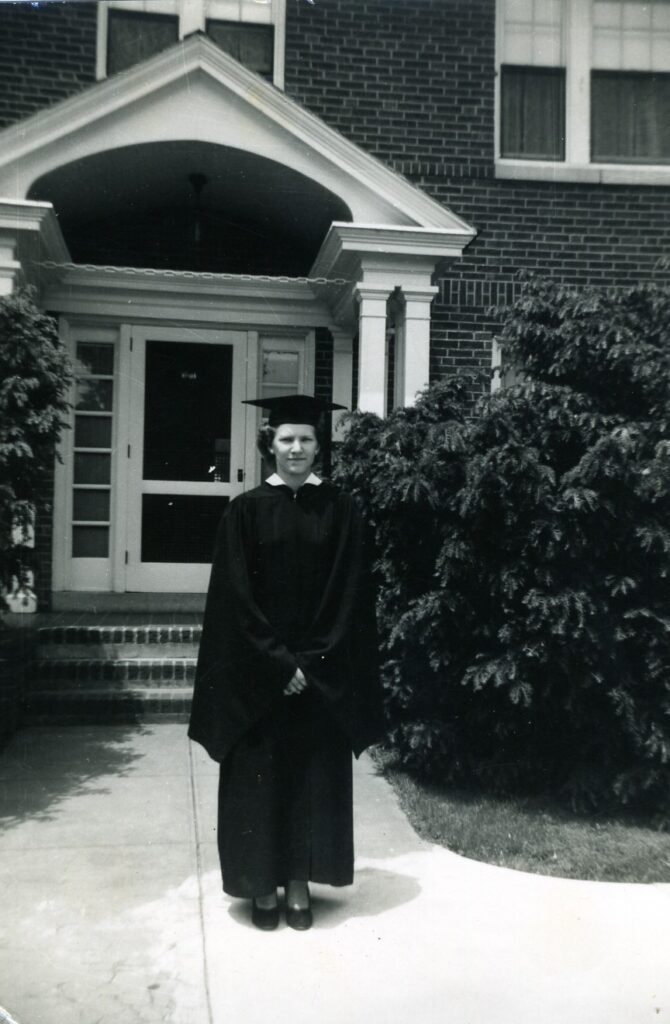 A black and white photo. A young woman stands in front of a brick building in graduation robes.