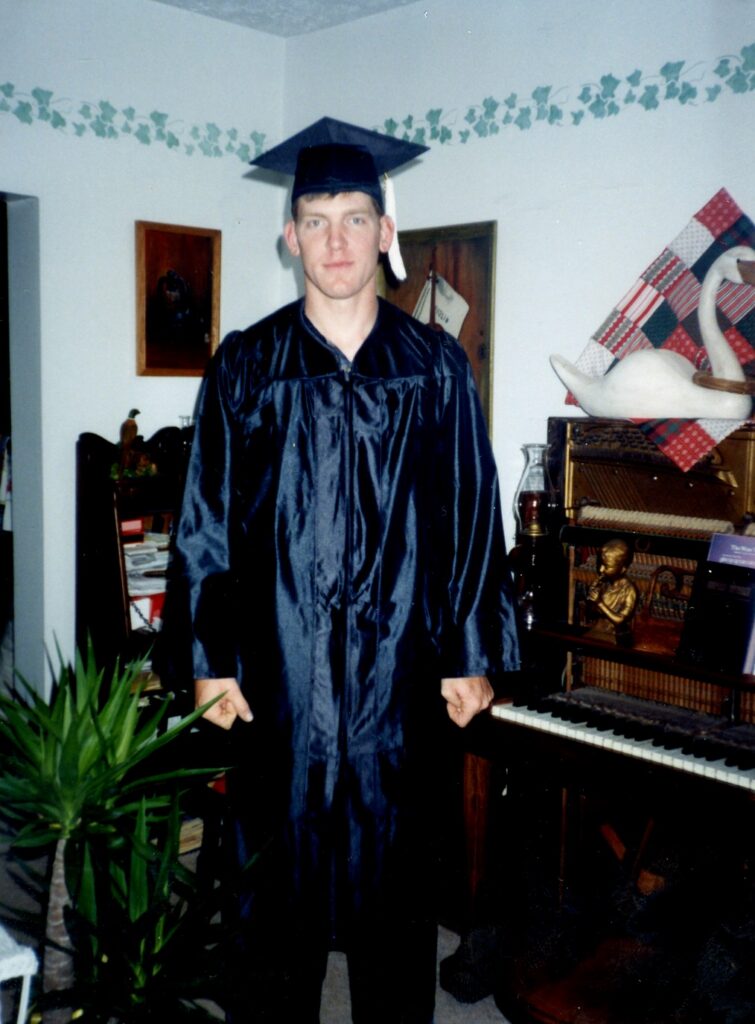 A color photograph, a young man stands in graduation robes.